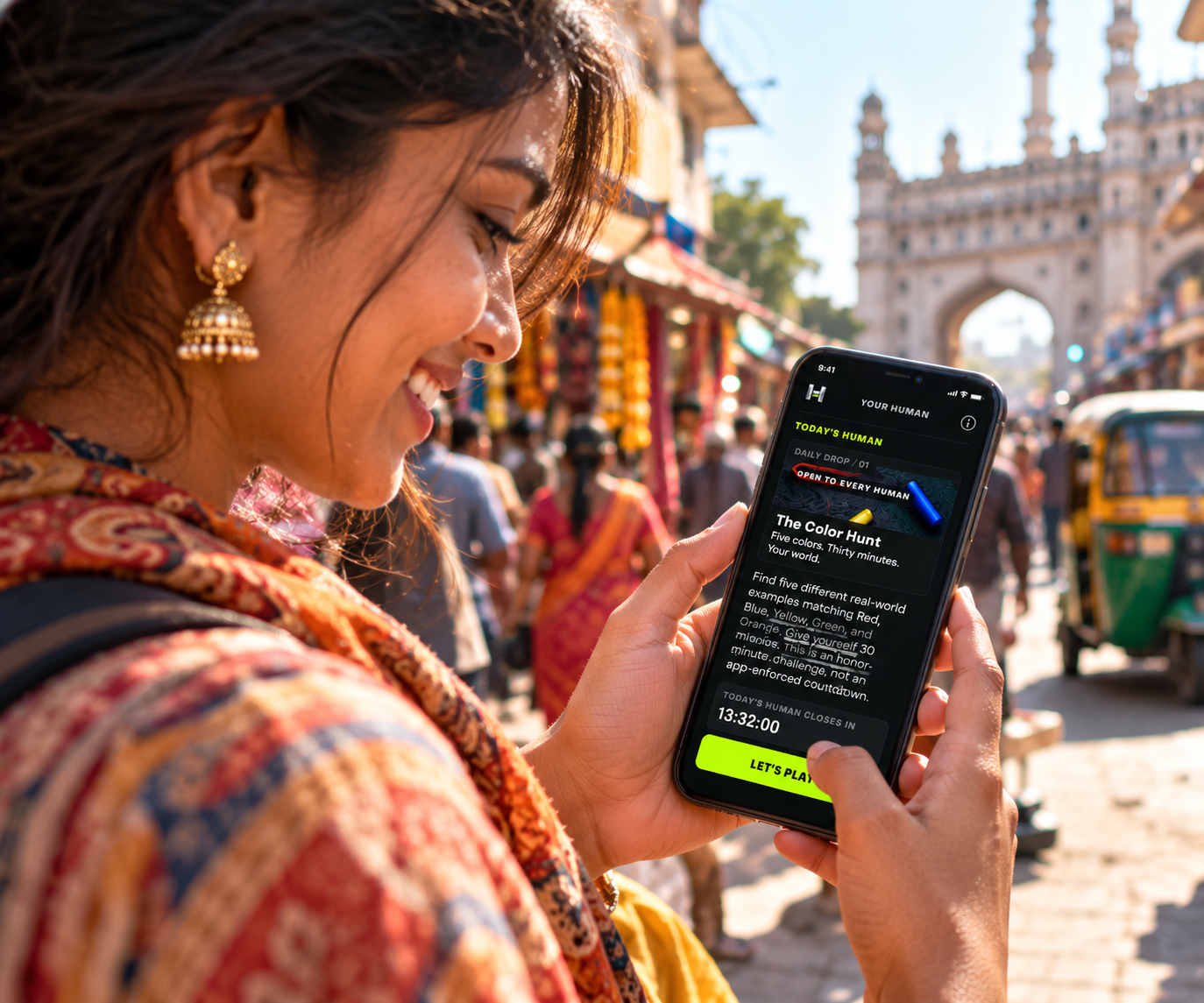 A smiling woman in a vibrant Indian market holds her phone showing today's HUMAN challenge