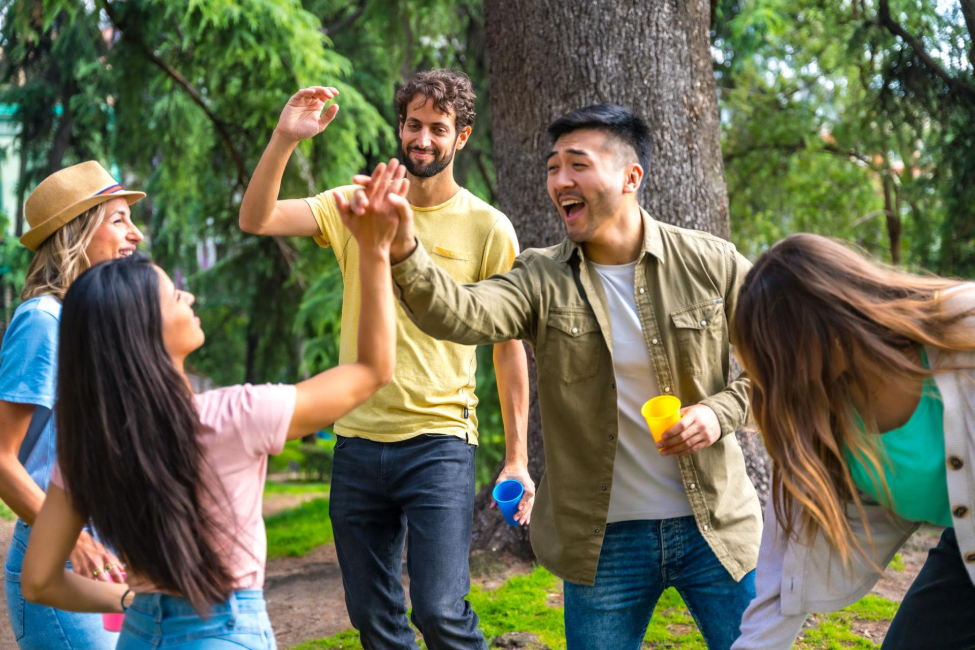 Friends in a park laugh and high-five while holding colourful cups