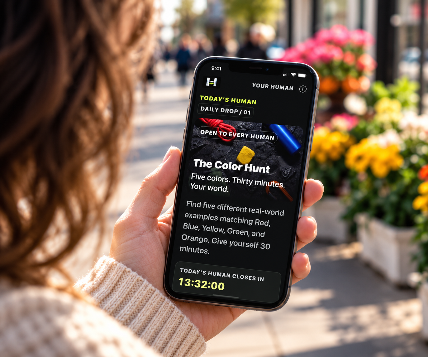 A woman on a sunny shopping street holds her phone showing The Color Hunt challenge beside a flower stand